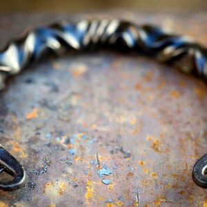 Puede incluir: Un tirador de metal negro forjado a mano con un dise&ntilde;o retorcido. El tirador tiene forma semicircular con extremos curvados, sobre una superficie oxidada y texturizada. El metal tiene un acabado oscuro y mate.