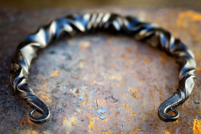 Puede incluir: Un tirador de metal negro forjado a mano con un dise&ntilde;o retorcido. El tirador tiene forma semicircular con extremos curvados, sobre una superficie oxidada y texturizada. El metal tiene un acabado oscuro y mate.
