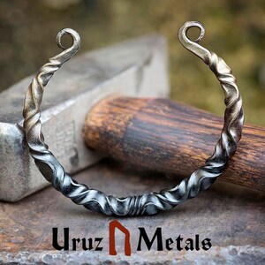 Puede incluir: Una pulsera de metal retorcido hecha a mano con un dise&ntilde;o en espiral. La pulsera tiene un acabado oscuro y envejecido con extremos rizados. El logotipo "Uruz Metals" es visible en la parte inferior de la imagen.