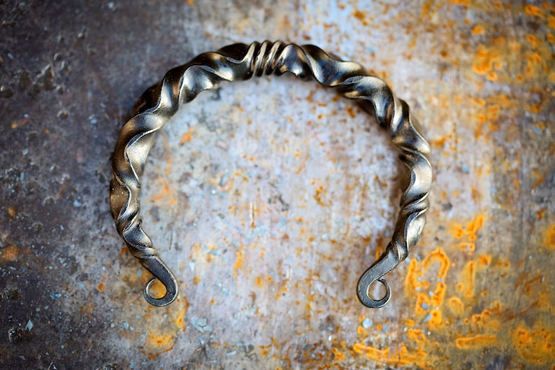 Puede incluir: Un brazalete de metal plateado hecho a mano con un dise&ntilde;o retorcido. El brazalete presenta un patr&oacute;n en espiral y extremos curvados. La joya se exhibe sobre una superficie texturizada y oxidada.