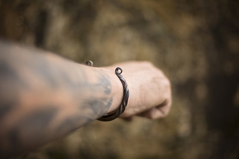 Puede incluir: Un brazalete de metal oscuro y retorcido con un dise&ntilde;o en espiral. El brazalete tiene un lazo en cada extremo. La imagen muestra el brazalete en el brazo de una persona con un fondo borroso.