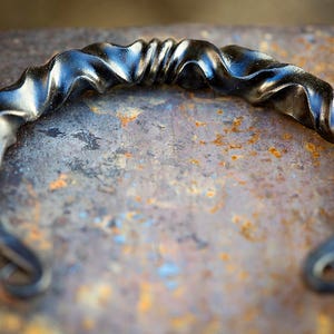 Puede incluir: Una pulsera de metal negro forjada a mano, con un dise&ntilde;o retorcido y ondulado. La pulsera tiene una forma curvada con extremos rizados. El metal tiene una superficie ligeramente reflectante, sobre un fondo texturizado y oxidado.