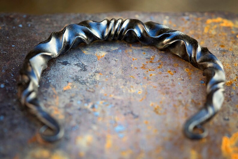 Puede incluir: Una pulsera de metal negro forjada a mano, con un dise&ntilde;o retorcido y ondulado. La pulsera tiene una forma curvada con extremos rizados. El metal tiene una superficie ligeramente reflectante, sobre un fondo texturizado y oxidado.