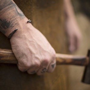 Puede incluir: Primer plano de una pulsera de pu&ntilde;o de metal oscuro con extremos puntiagudos en una mu&ntilde;eca. La persona sostiene un martillo con mango de madera. El fondo est&aacute; borroso, lo que sugiere un taller.