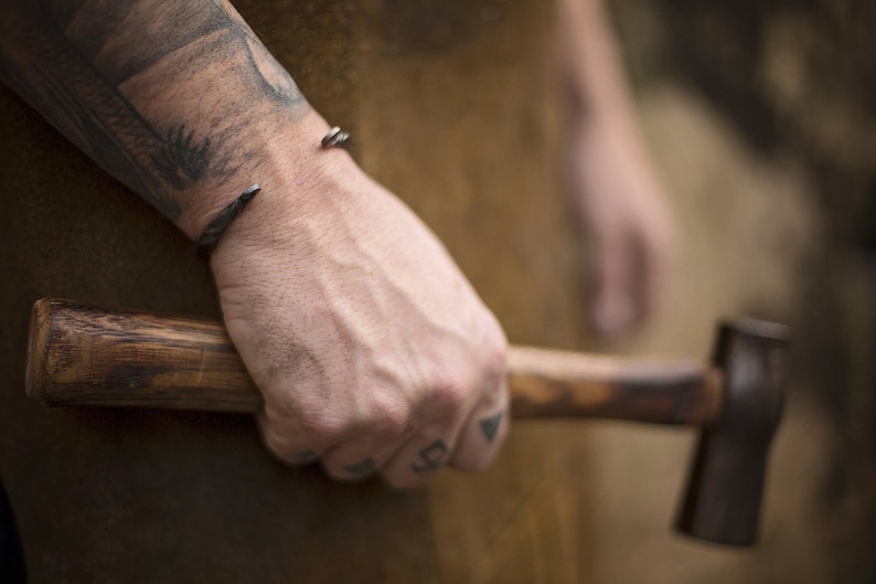 Puede incluir: Primer plano de una pulsera de pu&ntilde;o de metal oscuro con extremos puntiagudos en una mu&ntilde;eca. La persona sostiene un martillo con mango de madera. El fondo est&aacute; borroso, lo que sugiere un taller.