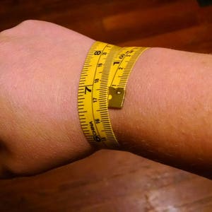 Puede incluir: Una cinta m&eacute;trica amarilla envuelta alrededor de una mu&ntilde;eca. La cinta muestra medidas en pulgadas, con una circunferencia de mu&ntilde;eca de aproximadamente 17,8 cm. La cinta est&aacute; hecha de un material flexible.