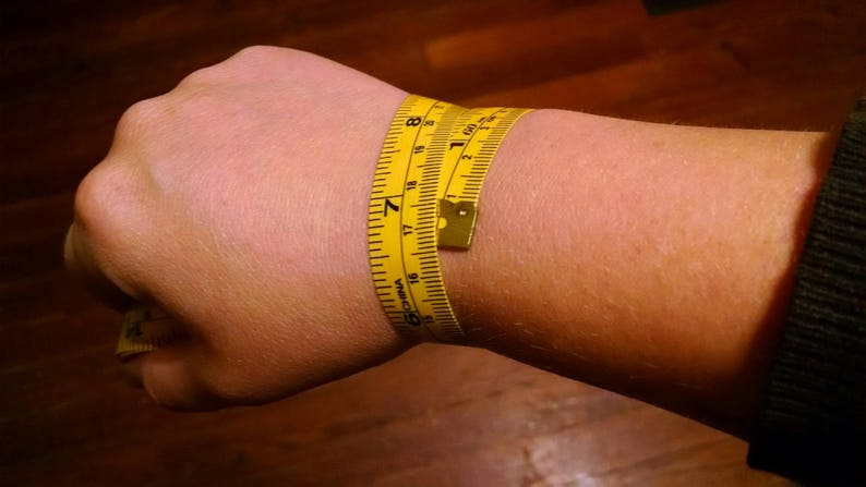 Puede incluir: Una cinta m&eacute;trica amarilla envuelta alrededor de una mu&ntilde;eca. La cinta muestra medidas en pulgadas, con una circunferencia de mu&ntilde;eca de aproximadamente 17,8 cm. La cinta est&aacute; hecha de un material flexible.