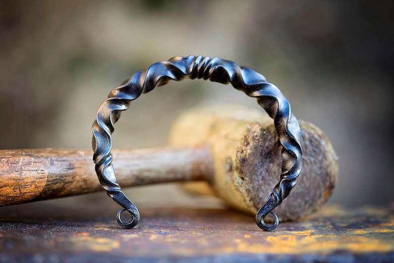 Puede incluir: Una pulsera artesanal de metal oscuro con un dise&ntilde;o retorcido. La pulsera est&aacute; hecha de hierro y tiene un patr&oacute;n en espiral. Los extremos de la pulsera tienen peque&ntilde;os bucles circulares. El fondo incluye un mazo de madera y una superficie met&aacute;lica.