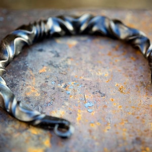 Puede incluir: Pulsera de metal retorcido y forjado a mano con un acabado oscuro y texturizado. La pulsera tiene un dise&ntilde;o en espiral y un cierre de gancho y ojo. La pieza de metal se muestra sobre una superficie oxidada y moteada.