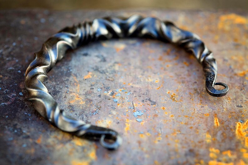 Puede incluir: Pulsera de metal retorcido y forjado a mano con un acabado oscuro y texturizado. La pulsera tiene un dise&ntilde;o en espiral y un cierre de gancho y ojo. La pieza de metal se muestra sobre una superficie oxidada y moteada.