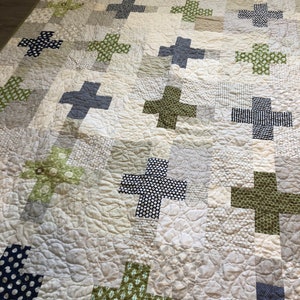 May include: A white quilt with a patchwork design featuring green, blue, and white plus signs. The quilt is quilted with a swirl pattern.