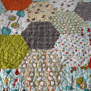 May include: A patchwork quilt with a hexagonal pattern. The quilt features various fabrics in shades of orange, green, blue, and brown. Some hexagons have patterned designs, including owls, trees, and geometric shapes. The quilt is quilted with a wavy stitch.