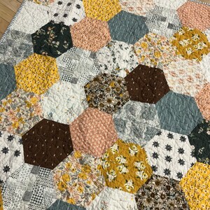 May include: A quilt made of various fabric scraps in shades of brown, yellow, white, and gray. The quilt is made up of hexagonal shapes, each with a different pattern. The quilt is quilted with a simple stitch pattern.