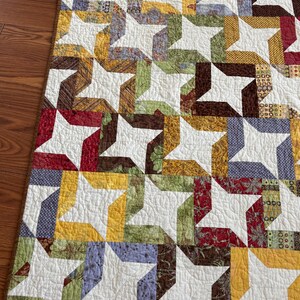 May include: A handmade quilt with a repeating star pattern. The stars are white, and the connecting pieces are various colors, including yellow, green, brown, red, and blue. The quilt is on a wooden surface.
