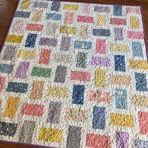 May include: A patchwork quilt with a white background and a variety of colorful rectangular patches in shades of pink, yellow, green, blue, and purple. The quilt is quilted with a simple stitch pattern.