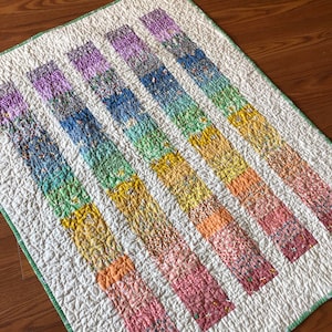 May include: A white quilt with seven colorful stripes of fabric. The stripes are arranged in a gradient pattern, starting with purple and ending with pink. The quilt is quilted with a simple stitch pattern.