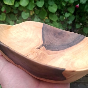 Könnte beinhalten: Handgeschnitzte Holzschale mit natürlicher, hellfarbiger Holzmaserung und einem dunklen, kontrastierenden Bereich. Die Schale hat eine organische, ovale Form und wird in einer Hand gehalten, mit einem verschwommenen grünen Hintergrund.