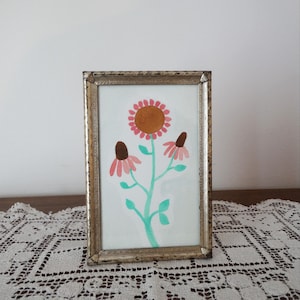 May include: A rectangular picture frame with a metallic, distressed gold finish containing a painting of a central flower with a brown centre and pink petals, flanked by two smaller flowers with brown centres and drooping pink petals on a stem.