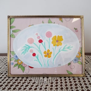May include: A framed watercolor painting featuring a bouquet of yellow and pink flowers with green stems and leaves, set against a light blue oval. The background is a pink floral pattern, all within a gold-colored frame.