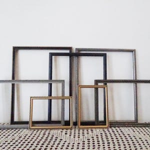 May include: A set of seven picture frames in various sizes and colours, including black, gold, and silver. The frames are arranged in a staggered pattern on a white surface.