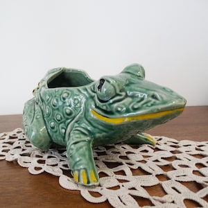 Peut inclure: Un cache-pot en céramique en forme de grenouille avec une finition verte brillante, présentant une surface texturée et bosselée, des yeux proéminents et une large bouche soulignée par une ligne peinte en jaune vif. Les pattes ont des bouts jaunes.