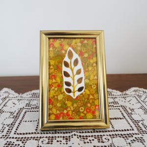 Puede incluir: Una obra de arte enmarcada con un marco de metal dorado. La obra presenta un diseño en forma de hoja blanca con detalles de hojas marrones, sobre un fondo floral en tonos de amarillo, verde y rojo. El marco es rectangular.