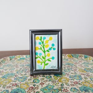 Op de afbeelding: Een kleine, ingelijste aquarel met een gestileerde boom met gele en turquoise bessen op een groene stengel. De schildering heeft een lichtblauwe achtergrond en is gesigneerd "AER". De lijst is zwart.