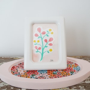 May include: A white rectangular picture frame with a watercolor floral print. The artwork features a teal stem with pink leaves and yellow dots. The frame sits on a pink oval tray with a floral fabric insert.