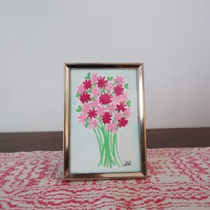 Puede incluir: Pintura enmarcada de un ramo de flores rosas y rojas con tallos verdes. La obra de arte está sobre un fondo azul claro y enmarcada en un marco de metal dorado. La pintura se exhibe sobre una superficie estampada en rojo y blanco.