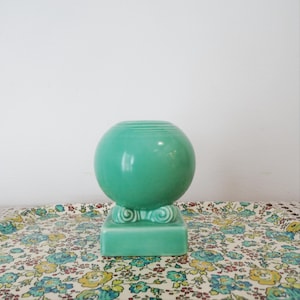 Op de afbeelding: Een bolvormige, mintgroene keramische vaas met een gladde, glanzende afwerking. De vaas heeft een ronde opening aan de bovenkant en rust op een vierkante voet met decoratieve, krulvormige details op de hoeken waar de bol de voet raakt.