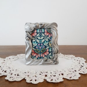 Puede incluir: Un marco de fotos plateado con un diseño floral. El marco tiene detalles decorativos de conchas marinas e hipocampos. La imagen en el interior muestra un colorido patrón floral con tonos rosas, azules y verdes.