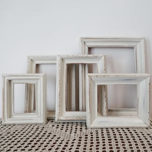 May include: A collection of four rectangular picture frames with a distressed white finish. The frames feature a stepped, beveled profile with visible wood grain and worn edges, arranged in a staggered overlapping formation against a plain wall.