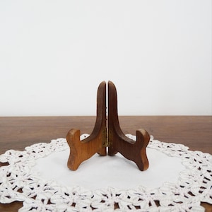 May include: A brown wooden display stand featuring two hinged pieces connected by a central brass-coloured metal hinge. The stand has a curved, upright design with two front-facing supports, constructed from smooth, polished wood with visible grain.