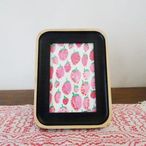 May include: A rectangular picture frame with a black border and a light wood frame. The artwork inside features a pattern of pink strawberries with green stems on a white background. The frame is resting on a red and white patterned surface.