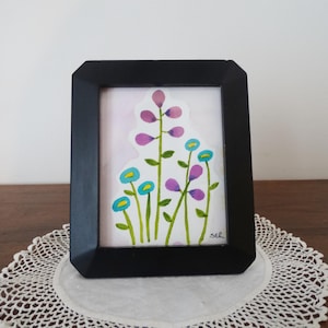 Könnte beinhalten: Ein gerahmtes Aquarellgemälde mit stilisierten Blumen in Lila, Blau und Gelb. Das Kunstwerk ist vor einem hellrosa Hintergrund platziert und in einem schwarzen, rechteckigen Rahmen gerahmt. Das Gemälde wird auf einer weißen Spitzendecke präsentiert.
