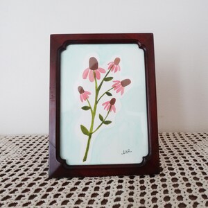 May include: A framed watercolor painting of pink coneflowers with brown centers and green stems and leaves. The artwork is set against a light blue background and is enclosed in a dark brown wooden frame.