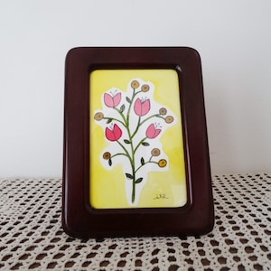 May include: A framed watercolor painting of a stylized floral arrangement. The artwork features pink tulip-like flowers and brown buds on a green stem against a yellow background. The frame is a dark, glossy brown.
