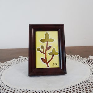 May include: A small, rectangular framed artwork with a plant design. The artwork has a yellow background with a brown stem and green leaves. The dark brown frame surrounds the artwork, which is signed "SER".