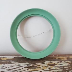 Puede incluir: Un marco decorativo redondo de color verde menta con un alambre fino para colgar. El marco se encuentra sobre un fondo blanco y descansa sobre una superficie de madera envejecida. El diseño circular del marco es simple y moderno.