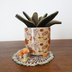 May include: A cylindrical ceramic planter with a light tan background featuring a repeating pattern of brown and orange mushrooms and chestnuts. The planter contains a dark green felt succulent plant and rests on three small orange feet.