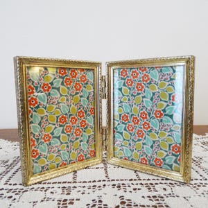 May include: A double picture frame in gold-tone metal. Each section displays a floral pattern with red flowers, green leaves, and yellow berries on a teal background. The frame has a decorative, textured edge.