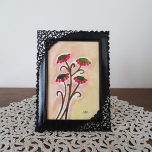 Könnte beinhalten: Gerahmtes Blumenbild mit einem schwarzen, verzierten Rahmen. Das Kunstwerk zeigt stilisierte Blumen mit roten und grünen Blüten auf braunen Stielen vor einem pfirsichfarbenen Hintergrund. Der Rahmen hat dekorative Eckakzente.