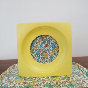 May include: A square, bright yellow picture frame with a circular opening. The frame holds a floral patterned fabric with blue, red, and yellow flowers. The frame is sitting on a floral patterned tray.