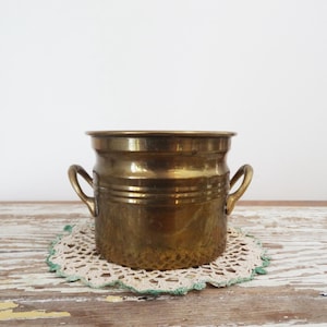 Peut inclure: Un pot en laiton vintage avec deux poignées incurvées. Le pot a une forme cylindrique avec des rainures horizontales et repose sur un napperon crème avec une bordure verte. Le fond est un mur blanc et une surface en bois vieilli.