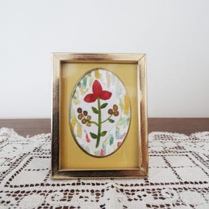Puede incluir: Un marco de fotos rectangular de metal dorado que contiene una pintura de acuarela de forma ovalada. La obra presenta una flor roja central con tallos verdes y bayas marrones, sobre un fondo de suaves pinceladas verticales multicolores.