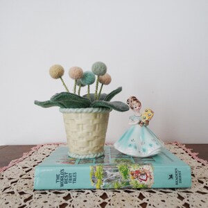 May include: A light yellow ceramic basket planter containing felted pom-pom flowers in muted tones of green, pink, and tan, positioned next to a porcelain figurine of a person in a blue dress holding a bouquet, all resting on a teal book titled The World's Best Fairy Tales.