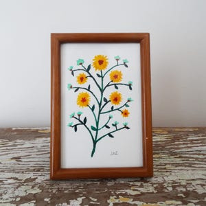 May include: A framed watercolor painting of a stylized floral design. The artwork features a central green stem with yellow flowers and small turquoise accents. The frame is made of brown wood, and the painting is on white paper.