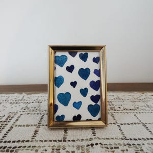 May include: A rectangular picture frame with a metallic gold finish and a beaded edge detail. The frame contains a white insert featuring a repeating pattern of hand-painted watercolor hearts in various shades of blue and navy.