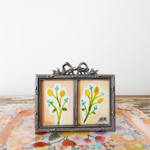 May include: A small, silver-toned double picture frame with a bow detail at the top. Each frame holds a watercolor painting of yellow and blue floral sprigs on a peach-colored background. The frame sits on a floral tray.