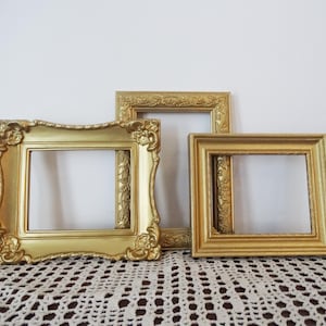 May include: Three ornate, gold-colored picture frames of varying sizes are arranged on a white surface. The frames have decorative edges and corners, with intricate floral and scrollwork details. The frames are empty, ready for photos or artwork.
